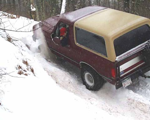 1988 Ford Bronco - A maroon and tan Ford Bronco navigates a snowy incline, kicking up snow as it goes.