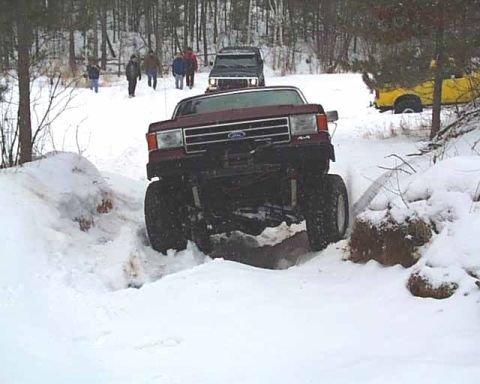 1988 Ford Bronco - A lifted red Ford Bronco navigates a snowy trail, with other vehicles and people in the background.