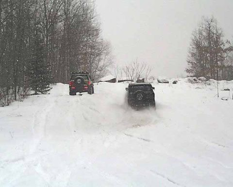 1988 Ford Bronco - Two SUVs navigate a snow-covered hill, with one kicking up a spray of snow as it drives.