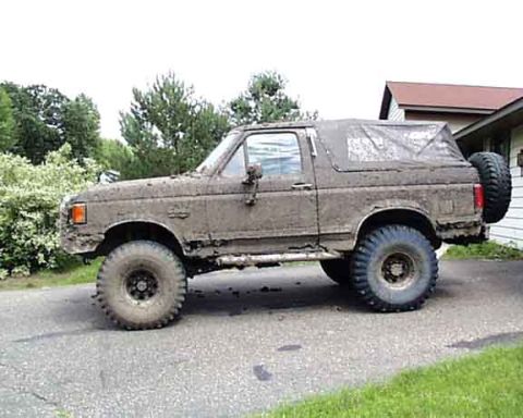 1988 Ford Bronco - This heavily muddied SUV, likely from the late 1980s, is parked on a driveway next to a house.