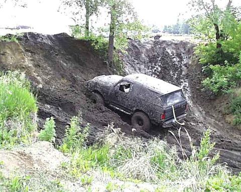 1988 Ford Bronco - This SUV is deeply stuck in mud on a steep embankment, with trees and greenery surrounding the scene.