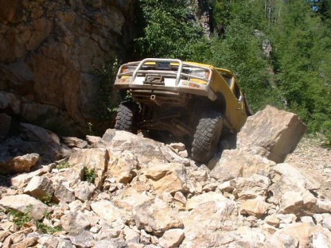 1988 Ford Bronco - A yellow 1988 Ford Bronco navigates a challenging rocky trail, its front bull bar visible as it climbs over large boulders.