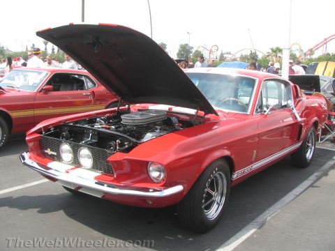 1968 Ford Mustang - A red 1968 Ford Mustang GT500 is displayed with its hood open, revealing the engine bay. Another classic Mustang is visible behind it.