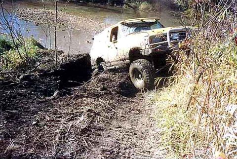 1988 Ford Bronco - A yellow off-road vehicle navigates a muddy trail beside a river.