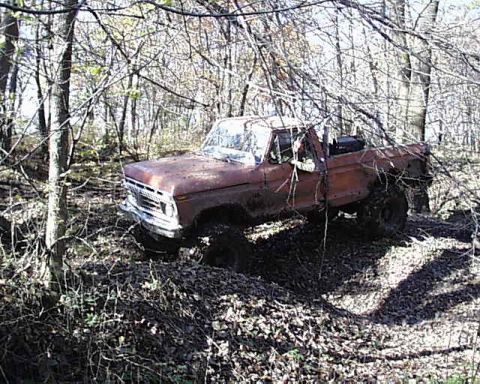 1988 Ford Bronco - An old pickup truck is partially submerged in mud and surrounded by trees and fallen leaves.