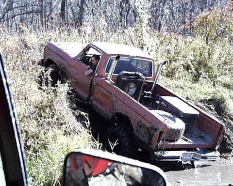 1988 Ford Bronco - An orange pickup truck is stuck in the mud, with a driver visible inside the cab and a spare tire in the truck bed.