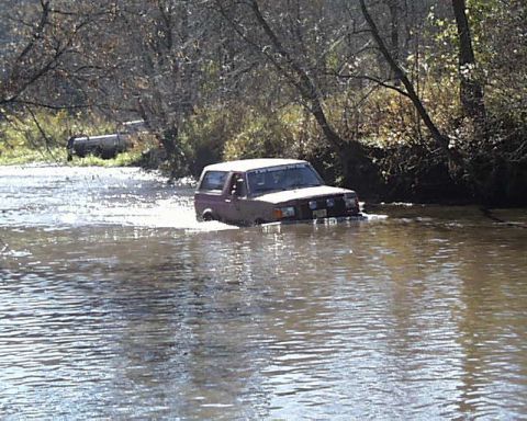 1988 Ford Bronco - A red 1988 Ford Bronco navigates through a shallow body of water with trees on the bank and another vehicle in the background.