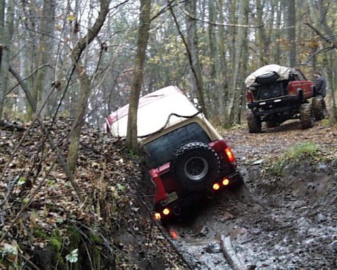 1988 Ford Bronco - Two off-road vehicles navigate a muddy trail in a wooded area, with one vehicle stuck in the mud.