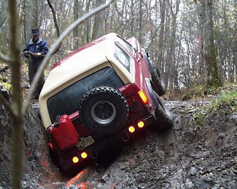 1988 Ford Bronco - A person watches as a red and tan SUV gets stuck in a muddy off-road trail.