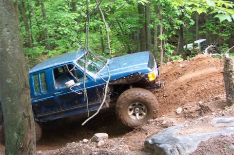 1988 Ford F-250 - A blue pickup truck navigates a steep dirt incline in a wooded area, with another vehicle visible in the background.