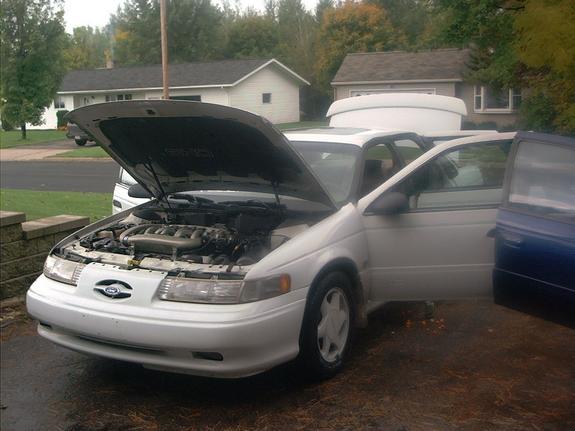 White Ford Taurus sedan with hood up showing engine and doors open in a wet driveway