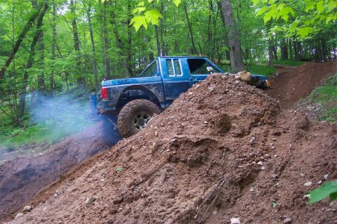 1988 Ford F-250 - A blue pickup truck navigates a steep, muddy incline in a wooded area, emitting a cloud of exhaust.