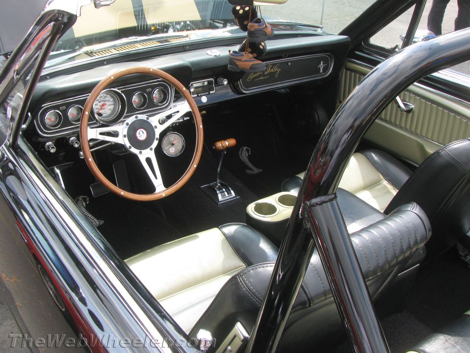 Black and white interior of a 1966 Ford Mustang convertible with a wooden steering wheel, roll bar, and Shelby Cobra badging