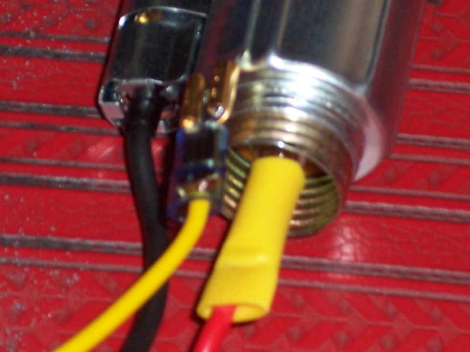 Close-up of a metal electrical connector with yellow and red wires attached, mounted on a red textured surface. A black wire is also visible