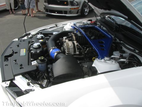 1968 Ford Mustang - The engine bay of a white 2008 Ford Mustang features a blue strut bar and a supercharger, with another Mustang visible in the background.