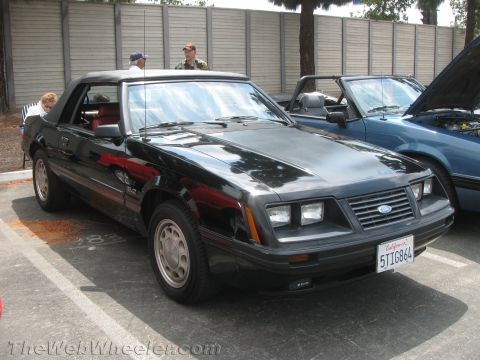 1968 Ford Mustang - This black convertible, likely from the early 1980s, is parked next to another car with its hood open.