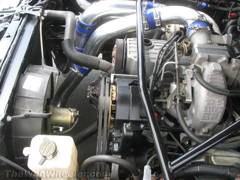 Engine bay showing turbocharger pipes, intercooler piping, alternator, and coolant reservoir. Text on intake reads '2.3 LITER EFI'