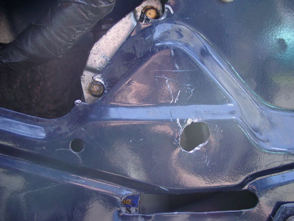 Close-up of dark blue vehicle panel with rough hole, scratches, and two bolts holding a metal bracket
