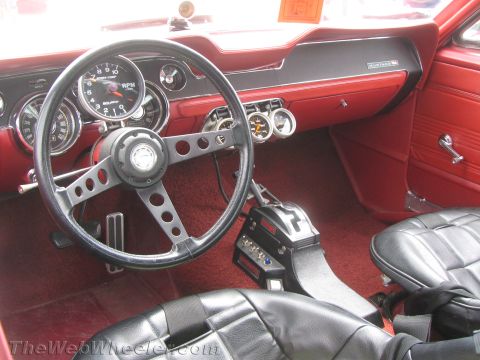 1968 Ford Mustang - The interior of a classic Ford Mustang shows a red dashboard, black racing seats, and a floor-mounted shifter.