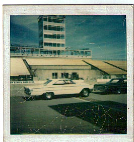 1963 Ford Galaxie - A white 1963 Ford Galaxie is seen at a racetrack, with a tower and grandstands in the background. Another dark vehicle is parked nearby.