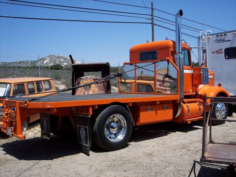 1981 Kenworth W900A - This orange flatbed truck features a prominent chrome exhaust stack and mud flaps with the Kenworth logo visible.