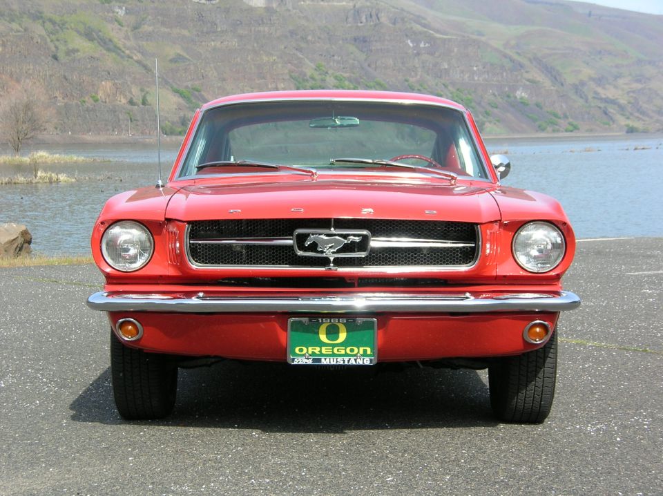 Red 1965 Ford Mustang hardtop front view with horse grille emblem and Oregon plate by a river