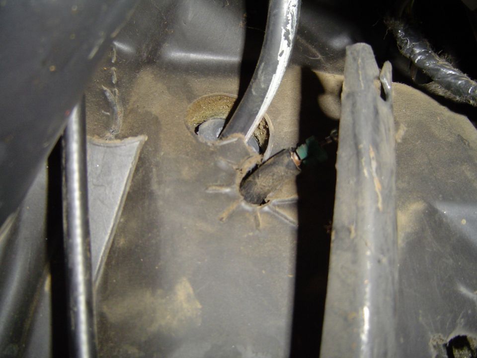 Cable routed through a grommet hole in a dusty metal vehicle panel, with a torn second hole and green connector