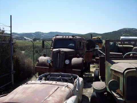 1981 Kenworth W900A - Several old, weathered vehicles sit in a lot under a clear blue sky, with rolling hills in the distance.
