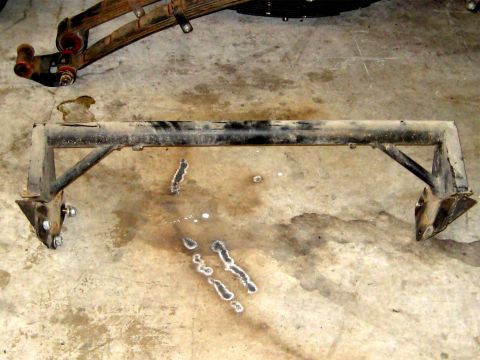 1996 Ford Bronco - A black metal suspension crossmember with welded brackets and leaf springs is shown on a concrete floor.