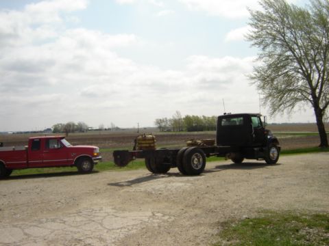 1992 Ford Bronco - A red pickup truck tows a black truck chassis on a dirt road with fields in the background.