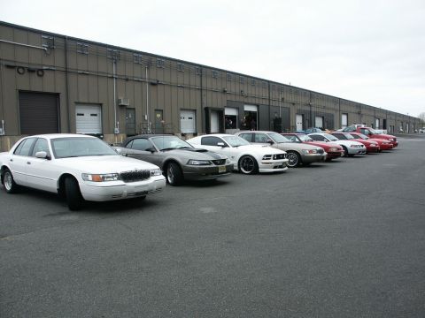 2001 Mercury Grand Marquis - Several cars, including a 2001 Mercury Grand Marquis, are parked in a line outside a large industrial building.