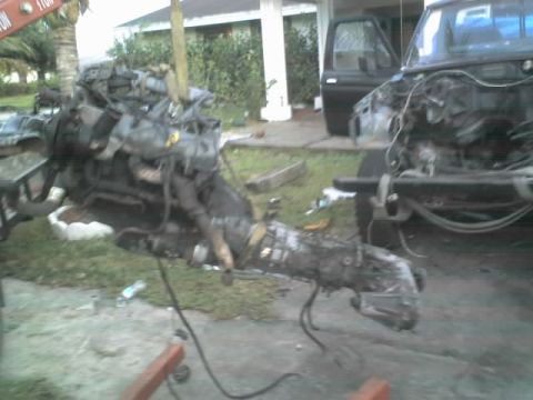 1995 Ford Bronco - The engine and transmission are being lifted out of a black truck, revealing the truck's partially disassembled front end.
