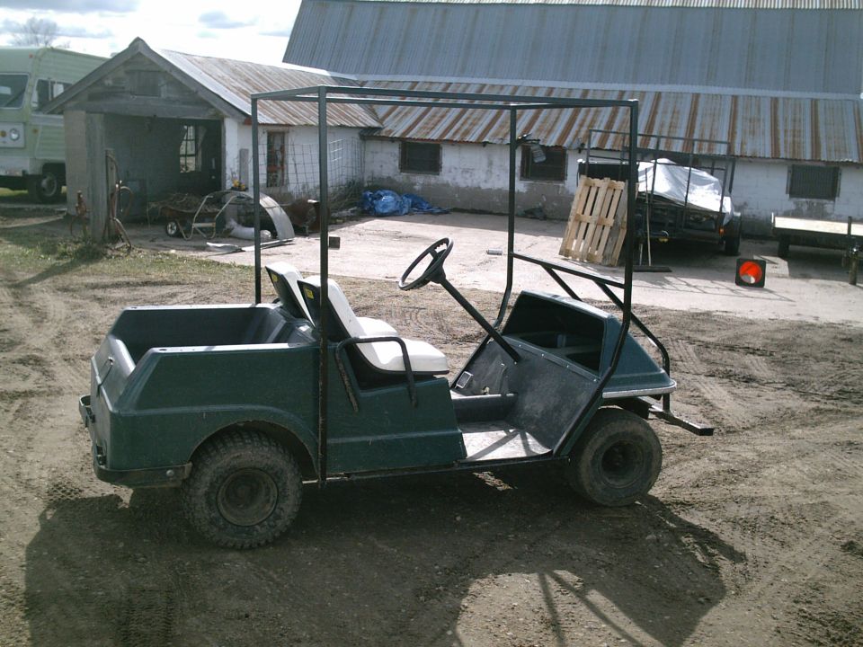 Green golf cart with black roll-cage frame, side view on dirt lot by farm buildings