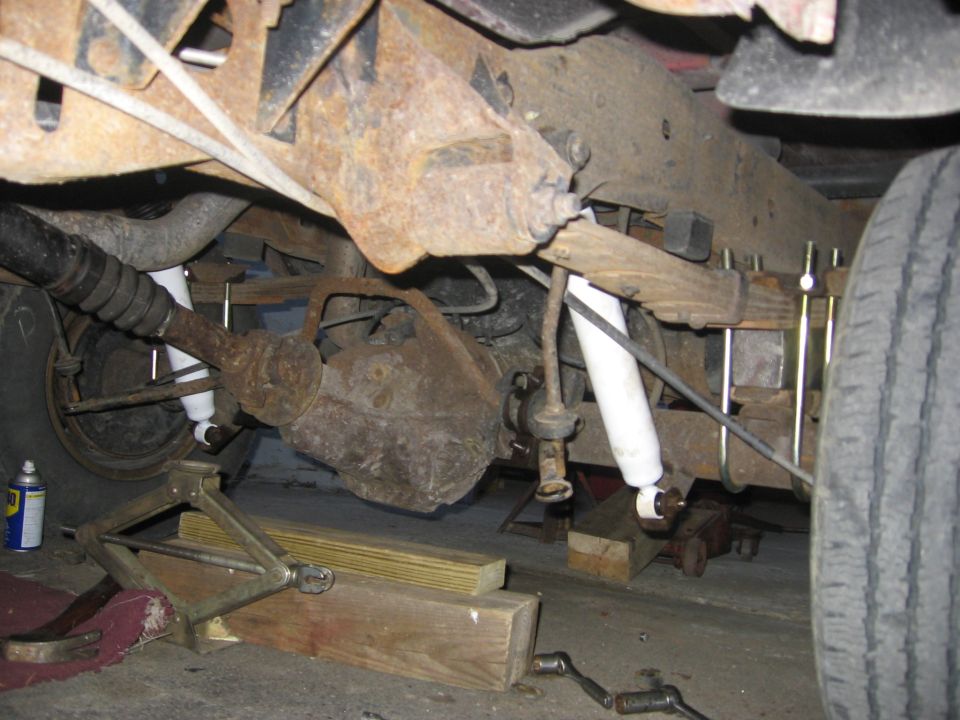 Underneath view of rusty rear axle, leaf springs, and shocks. A jack and wooden blocks are visible