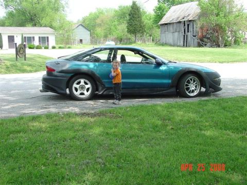1993 Ford Probe - A child stands beside a modified teal sports car parked on a street with a barn in the background.