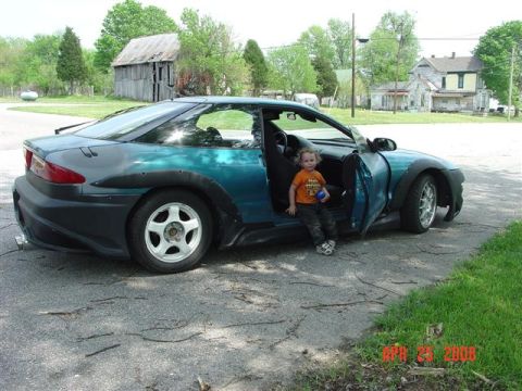 1993 Ford Probe - A child sits in the open driver's door of a modified teal and black coupe parked on an asphalt surface.
