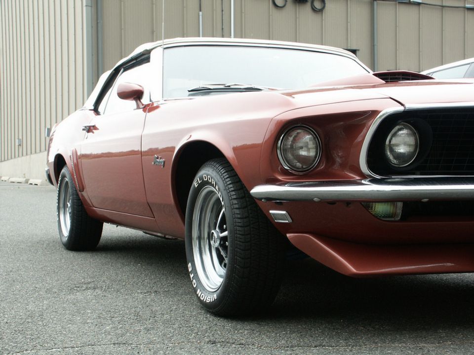 Front 3/4 view of a brown 1969 Ford Mustang convertible with a white top. It has a scoop on the hood and classic wheels
