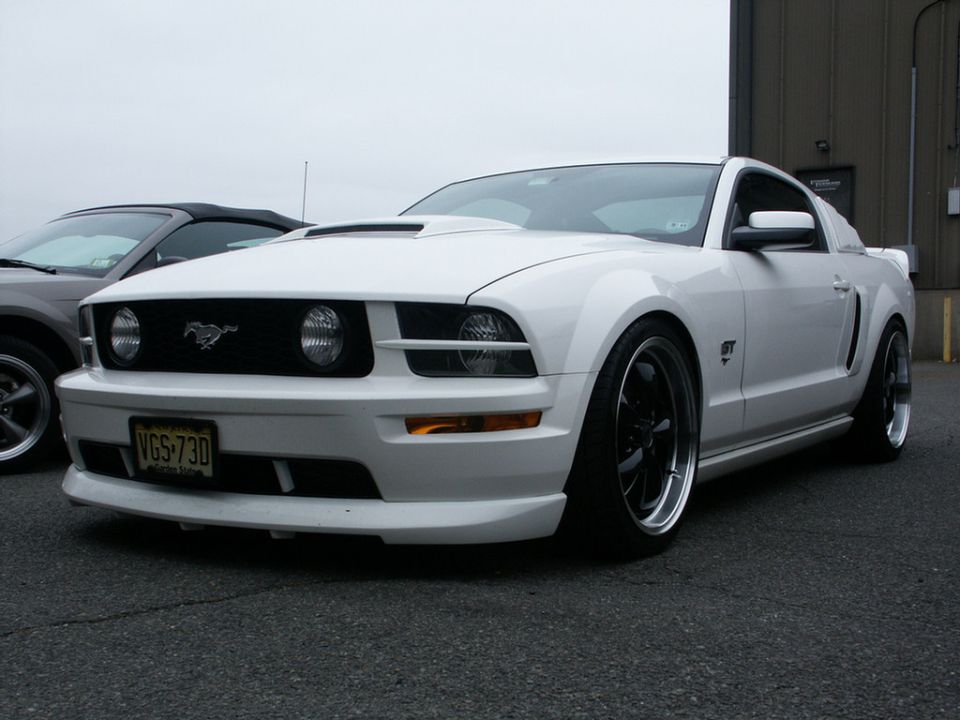 White 2005 Ford Mustang GT with aftermarket wheels and hood scoop, parked next to a grey convertible