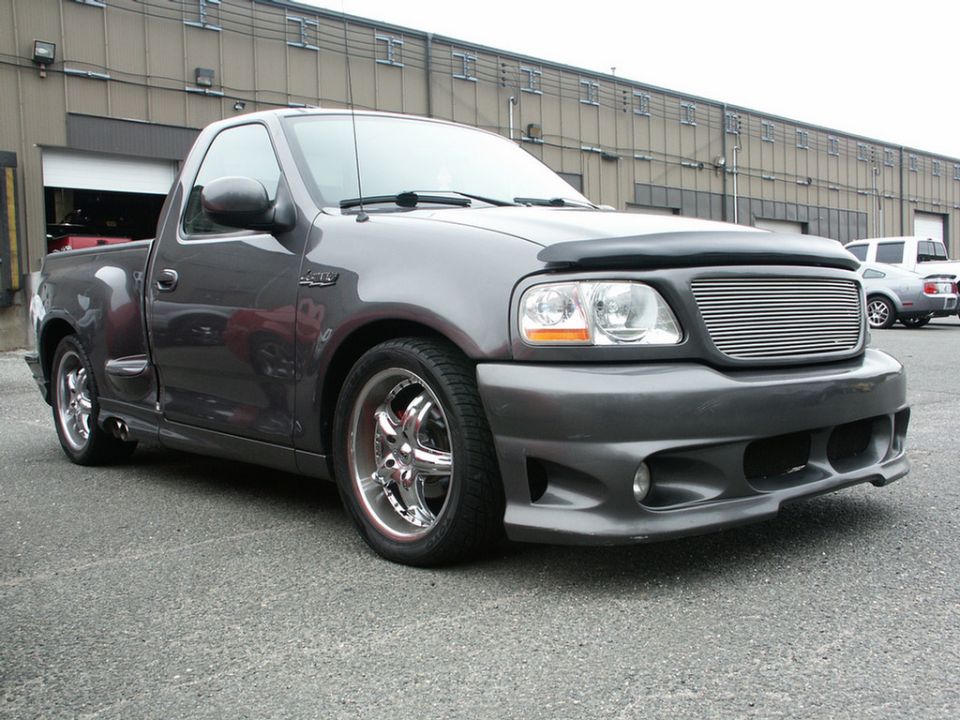 Gray Ford F-150 Lightning pickup truck with aftermarket grille and wheels parked outside a building. Another car is visible in the background