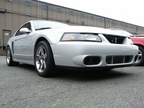 2001 Mercury Grand Marquis - A silver Ford Mustang is parked in front of a commercial building, with a red vehicle visible to its right.