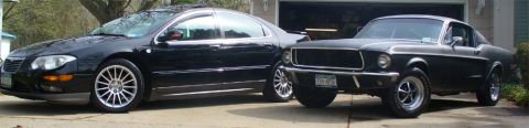 2002 Chrysler 300M - A classic black Ford Mustang is parked next to a modern black Chrysler 300M in front of a garage.