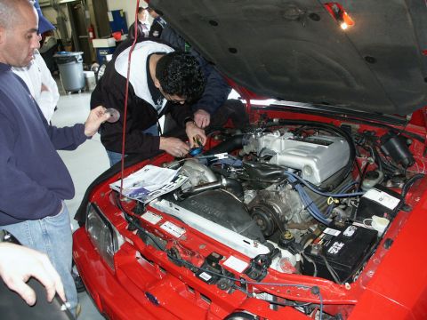 2001 Mercury Grand Marquis - Mechanics gather around a red car's engine compartment, appearing to perform maintenance or diagnostics.