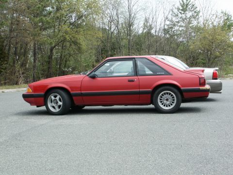 2001 Mercury Grand Marquis - A red coupe, likely a 1987 Ford Mustang, is parked beside a silver sedan on an overcast day.