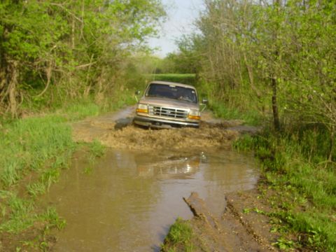 1996 Ford Bronco - Head-on view of a tan/gold 1990s Ford Bronco pushing through a long muddy water hole on a tree-lined two-track trail, throwing a brown bow…