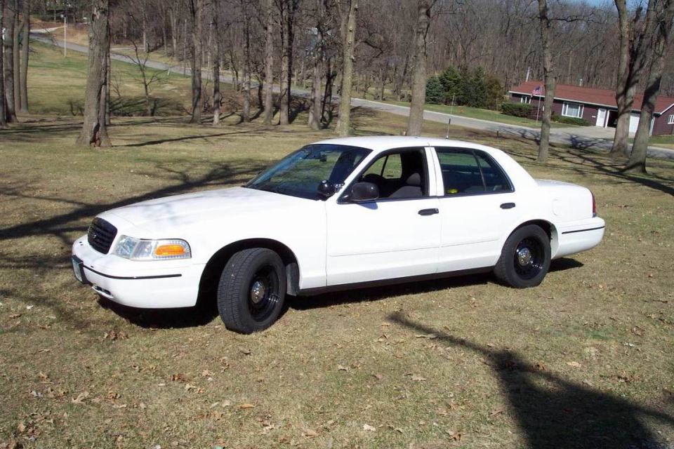 White Ford Crown Victoria sedan parked on a leaf-covered lawn in front of bare woods
