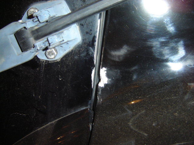 Close-up of a black car door with a damaged hinge and chipped paint near the seam. A Supermotors.net watermark is visible
