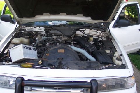 1996 Ford Crown Victoria - The engine bay of a white 1996 Ford Crown Victoria is visible with the hood open, revealing the engine and an Interstate battery.