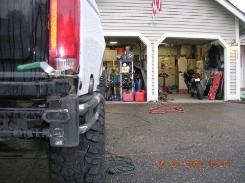 1996 Ford Bronco - A close-up view of a truck's rear tire and bumper, with a garage and motorcycle in the background.
