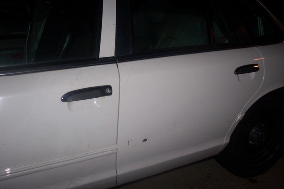 Side view of a white sedan's doors and rear wheel. There are two door handles and a small dent on the lower door panel
