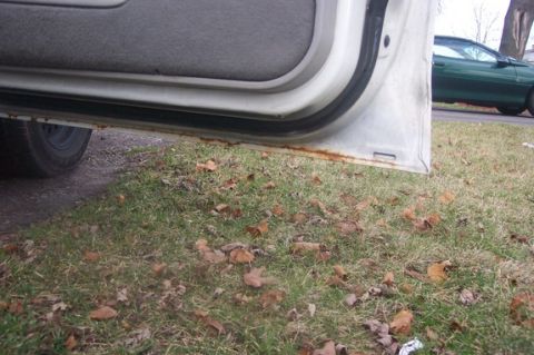 1996 Ford Crown Victoria - The lower part of an open car door reveals rust on the sill, with a green car in the background.